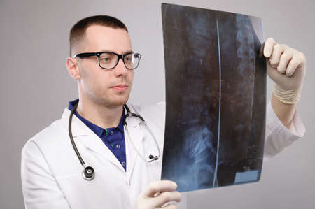 A young male doctor in glasses with a stethoscope around his neck holds an X-ray image of the spine...