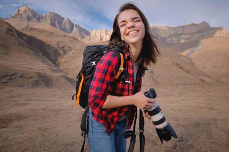 Professional photographer young attractive woman with a backpack and a professional camera in her hands laughs looking at the camera. against the backdrop of epic mountains at sunsetの写真素材