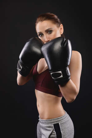 Caucasian fitness girl in boxing gloves stands in a rack on a black background, portrait of a strong and independent woman fighterの写真素材