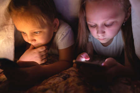 Two Child watching video smartphone under the blanket on the bed at night when light flashes bounce off the screen, children use addiction games and cartoon conceptの写真素材