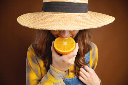 A young attractive Caucasian woman with long hair, wearing a denim overalls and a straw hat that covers her face. holding a cut orange in his hands bites him.の写真素材