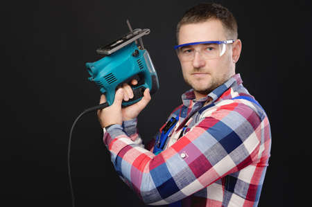 Caucasian man builder carpenter in a shirt and overalls with an electric tool in his hands. Studio portrait of friendly artisan businessmanの写真素材