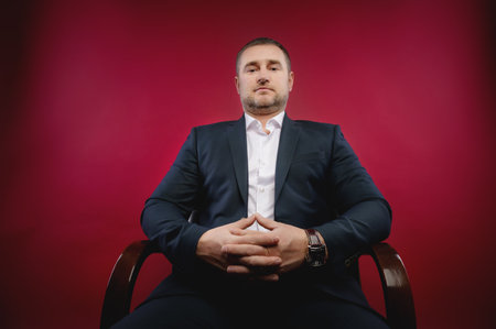 Business portrait of an attractive middle-aged man with stubble on his face in a black suit. Studio photo. The boss sits and looks down at the cameraの写真素材