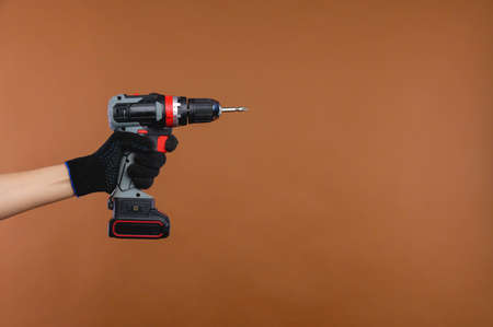 Female hand in a working rag glove holds an electric screwdriver on a battery on a brown background
