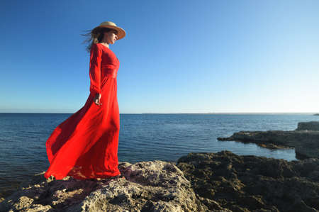 Lovely young slender caucasian woman in a silk pancake dress and a straw hat stands in a picturesque boroco pose on the rocky ocean coast on a sunny morningの写真素材