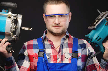 Caucasian man builder carpenter in a shirt and overalls with an electric tool in his hands. Studio portrait of friendly artisan businessmanの写真素材