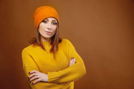 Portrait of a young attractive Caucasian woman in a yellow sweater and orange hat on a brown background. Winter clothing and seasonal insulationの写真素材