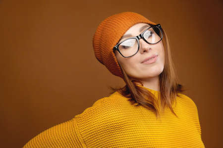 Portrait of a young attractive Caucasian woman in a yellow sweater and orange hat glasses on a brown background. Winter clothing and seasonal insulationの写真素材