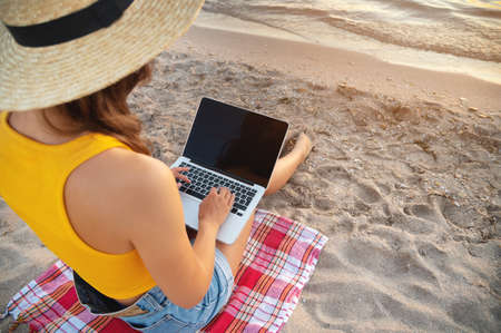 A slender young lady in denim shorts, a yellow tank top and a straw hat sits on a blanket on the sandy seashore and works on her laptop at sunset.の写真素材