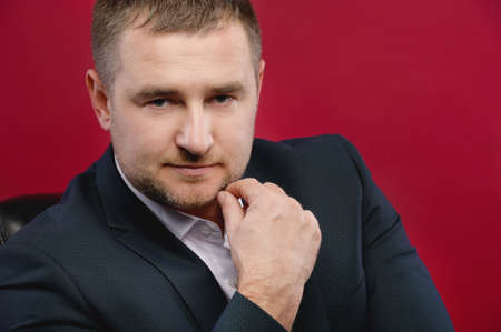 Close-up Business portrait of an attractive middle-aged man with stubble on his face in a black suit. Studio photo. The boss sits and looks down at the cameraの写真素材