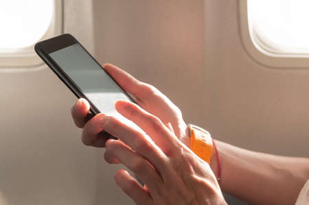 womans hands holding a black smart phone with blank desktop screen next in the cabin at the window airplaneの写真素材