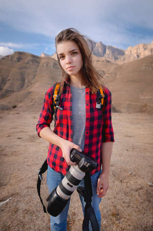 girl stands in a shirt a camera in her hand against a background of mountains
