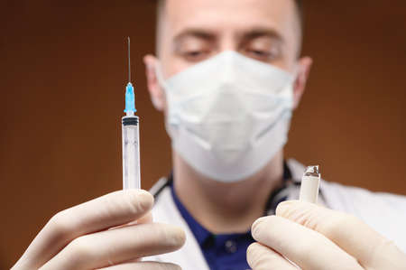 A young Caucasian doctor holds a syringe with an injection in his hand and an open ampoule with an...