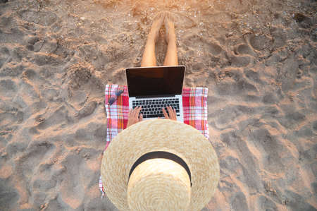 Young caucasian woman freelancer in a thatched sits on a sandy beach by the sea at sunset with a laptop and performs remote work or internet surfing. View from aboveの写真素材