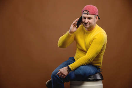 Studio Portrait of a plump caucasian man in a yellow sweater and cap talking on a cell phoneの写真素材
