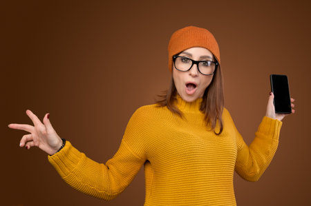 Shocked Caucasian young woman in yellow sweater and hat with phone in hand on brown background. Shock surprise gesturesの写真素材
