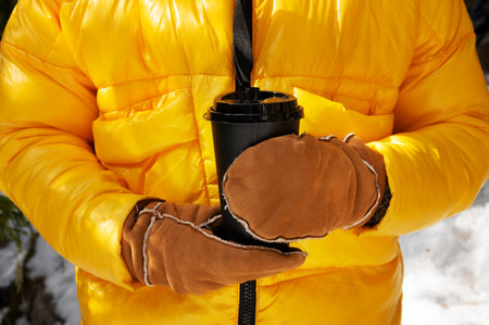 A woman dressed in a yellow down jacket and mittens holds a paper cup of hot coffee or tea in her...