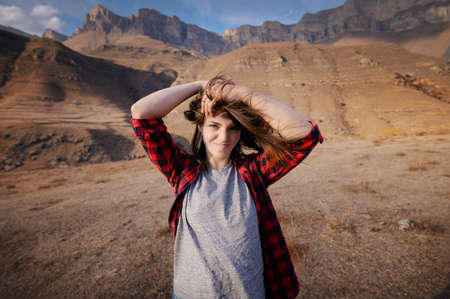 happy attractive hipster traveler girl with windy hair and smiling against the backdrop of sunny mountains copy space. travel and wanderlust. Hands in hair look at the cameraの写真素材
