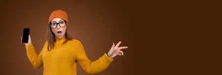 panoramic Shocked Caucasian young woman in yellow sweater and hat with phone in hand on brown...