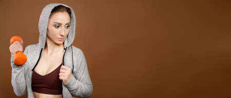 panoramic Portrait of a strong Caucasian athletic woman in a tracksuit with a hood holds a fitness dumbbell in her hand and looks to the side. Sports lifestyle. Studio shotの写真素材