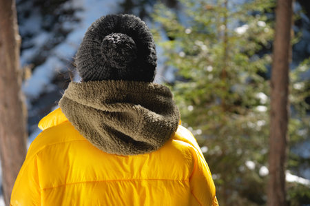 back portrait, a girl in a knitted hat and a yellow jacket stands in the forest and looks at the...