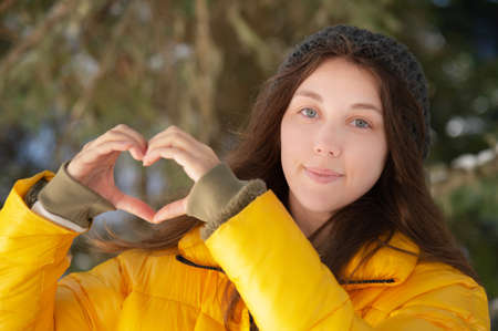 Attractive young Caucasian woman in a down jacket and hat shows a gesture of heart and love to the camera with her hands. Against the background of a coniferous winter forestの写真素材