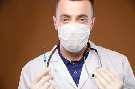 A young Caucasian doctor holds a syringe with an injection in his hand and an open ampoule with an...