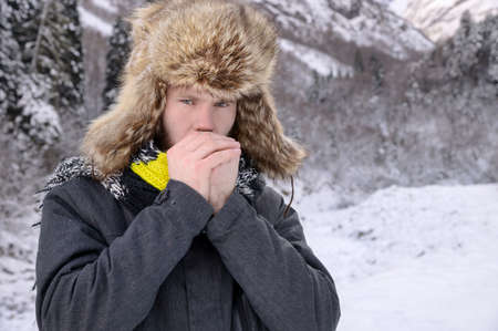 Portrait of a Caucasian man in a large fur hat and knitted scarf, shivering in the cold winter, breathing and rubbing his hands. Man in winter forest surrounded by snowの写真素材