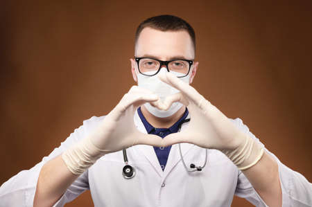 Portrait of young male doctor in glasses holding heart gesture in studio on gray background