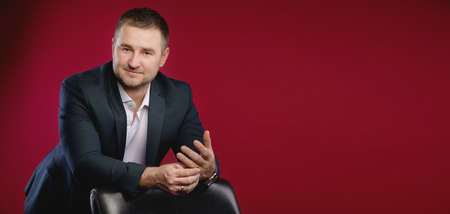 panoramic Portrait of an attractive smiling Caucasian male businessman in a suit. Stands in the studio on a red background, leaning on a leather armchair and holding on to the ring fingerの写真素材