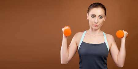 Panoramic shot of an athletic caucasian woman with dumbbells in her hands. Stands on a brown background with dumbbells in his hands and looks at the cameraの写真素材