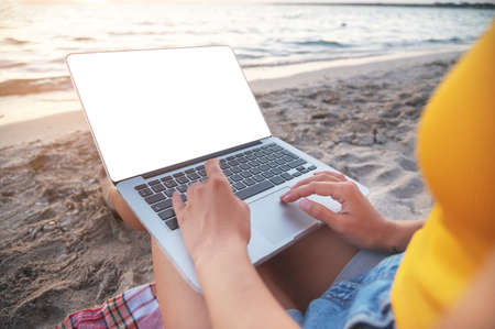 Close-up of a cut-out laptop screen on the lap of a young caucasian woman sitting on a sandy beach by the sea at sunset. Freelance and remote work presentation copy spaceの写真素材