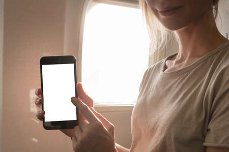Mockup image of a woman holding and looking at smart phone with blank white screen next to an airplane window with clouds and sky backgroundの写真素材