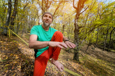 Portrait of a mature man in a scarf on his head sits on a taut slackline explains how to balance in...