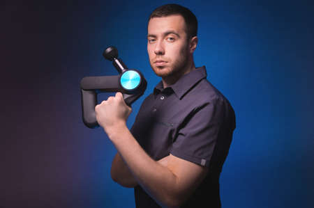 Portrait of a confident professional masseur with a percussion massager in his hands. Low key, blue...