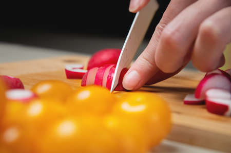 Close-up of female hands cut fresh raw radish on a wooden board against the background of yellow...