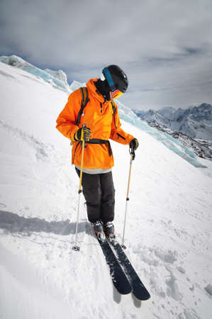 A professional skier athlete in a red jacket, helmet and mask stands on a slope against the backdrop...