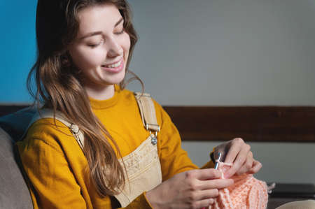 Young happy caucasian woman smiling sitting on sofa and crocheting wool product. Womens hobby production of clothes from woolの写真素材
