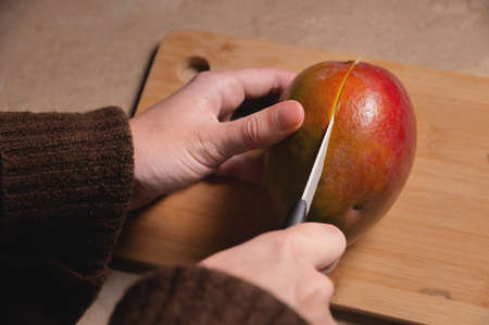 Close-up of womans hands cutting fresh mango on wooden cutting board at home kitchenの写真素材