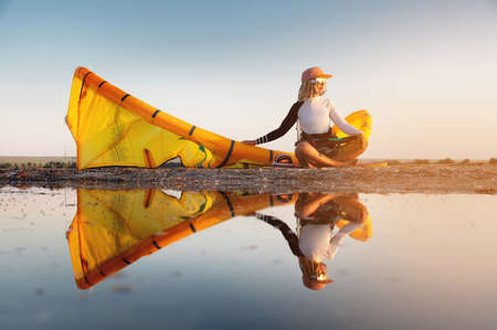 Attractive stylish young caucasian woman in cap sunglasses and kitesurfer outfit sits on the sandy shore next to her kite at sunset reflecting in the waterの写真素材
