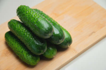 A pile of fresh small green cucumbers on a wooden cutting board on a white tableの写真素材