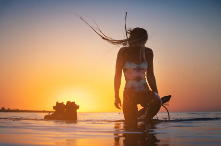 Silhouette frame. A young slim sexy female kitesurfer with a plank and a kiteboard stands in the water in the shallow water at sunset. Water sports. Stylized frameの写真素材