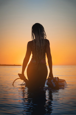 Silhouette frame. A young slim sexy female kitesurfer with a plank and a kiteboard stands in the water in the shallow water at sunset. Water sports. Stylized frameの写真素材