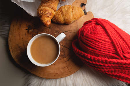 Atmospheric still life white mug of coffee next to fresh croissants in a paper bag next to red yarn. Home comfort in soft focusの写真素材
