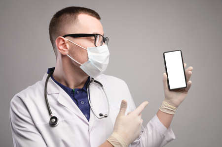male doctor in a coat, mask, glasses, gloves and with a stethoscope holding a smartphone in his hands, isolated screen, studio shooting Pointing at the screenの写真素材