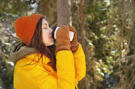 Girl in casual clothes with a glass of hot drink in her hands. Young woman in winter forest or park....