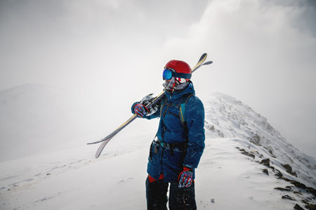 The skier holds a pair of skis and looks at the snow-capped mountains. A guy with skis on his...