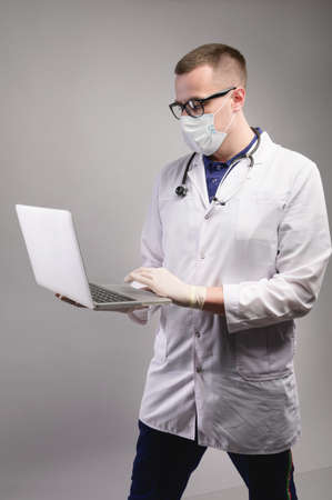 Young caucasian male doctor in uniform with a laptop for notes, on a gray background. Physician-therapist medical student. Medical education conceptの写真素材