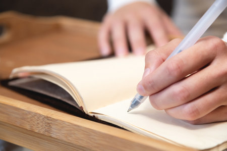 Close-up of a young woman in overalls and a sweater writing with a black gel pen in a leather notepad on a wooden tray table while sitting on the floor by the sofa at homeの写真素材