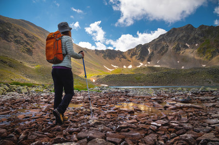 a stylishly dressed woman goes with trekking poles and a backpack to a mountain lake. hiking in the...